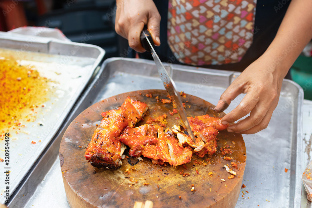 Street vendor cutting a grilled chicken marinated with curcuma on wooden cutting board; Thai street food market. 