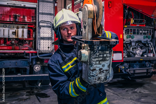 A fireman with uniform and helmet holding a chainsaw with fire truck in the background