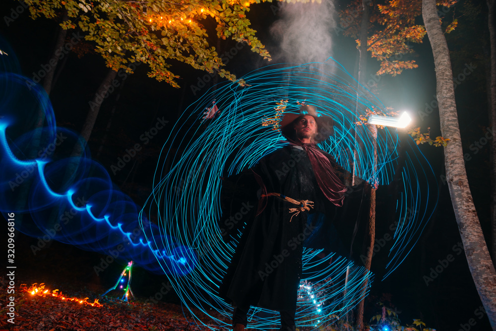 Fototapeta premium Sorcerer (magician) casting a spell while standing in the magical woods. Holding his magical staff. Magic illumination. Autumn holiday celebration. Mystery and nightmare concept.