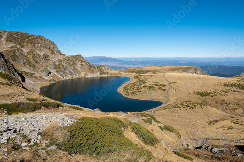 Blue glacial lake with steep rocky banks, the Kidney Lake, part of The Seven Rila Lakes, and a clear blue sky in the background. Around it can be seen several hiking trails, Rila mountain, Bulgaria 