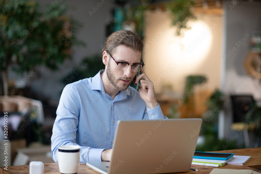 Young business man working on laptop talking on smartphone. Young business man working on laptop talking on smartphone.