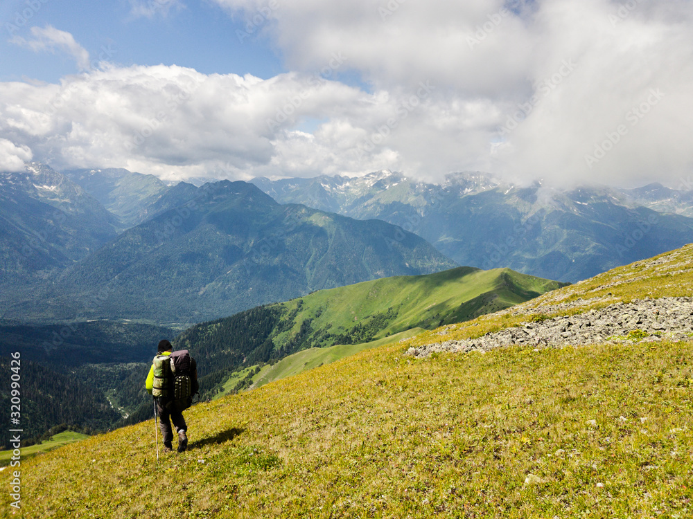Hiker with a backpack goes on the grassy slope on a background of mountains. Back view. Beautiful sunny day in Arkhyz. Concept of healthy lifestyle, trekking activity, hiking adventure