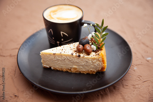 A black cup with a coffee is on a black plate near a piece of cake on which there are nuts and fruits