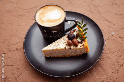 A black cup with a coffee is on a black plate near a piece of cake on which there are nuts and fruits