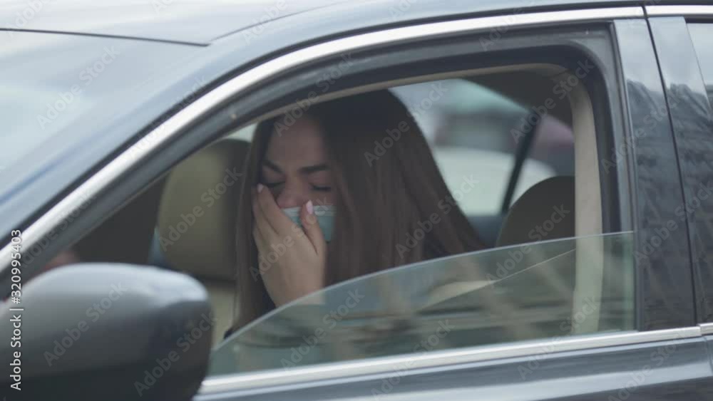 Side view portrait of young woman in protective mask sitting in car and coughing. Young woman with symptoms of disease sitting on driver's seat. Coronavirus, virus, global hazard. Side view portrait of young woman in protective mask sitting in car and coughing. Young woman with symptoms of disease sitting on driver's seat. Coronavirus, virus, global hazard.