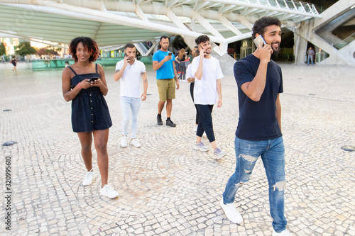 Group of young people using smartphones while walking on street. Cheerful friends strolling on city square and using digital devices. Communication, technology concept