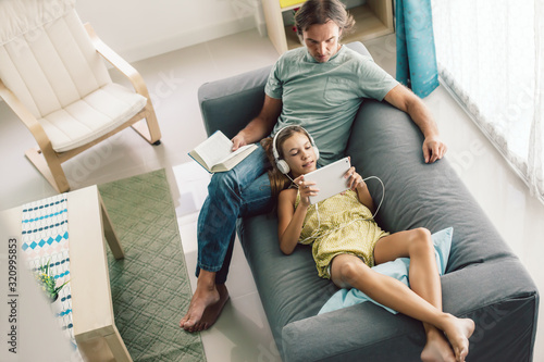 Father and pre teen daughter playing tablet and reading book while relaxing together on couch in room at home