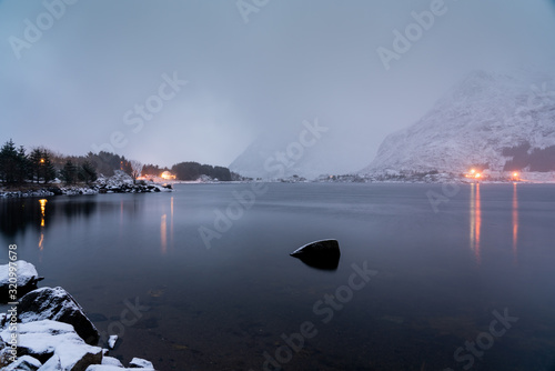 Winter landscape with a lake near mountains covered with snow and small village with evening lights.