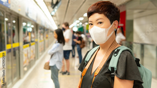 Social distancing, Beautiful middle aged Asian female traveller wear medical face mask to protect from infection of virus, pandemic of Covid-19 transmission in crowded underground train platform.