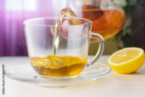 Hot herbal tea from a teapot is poured into a transparent cup. Nearby lies a lemon. The concept of healthy organic drinks.