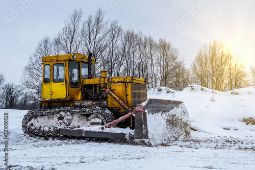 Yellow excavator on new construction site, with the bright sun and nice blue sky in the background. Yellow bulldozer in the winter on the street, snowdrifts and snow slopes