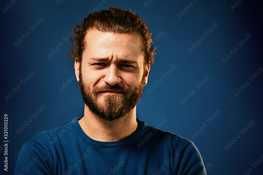Young attractive caucasian bearded hipster standing in front of blue background, frowning and looking ar camera. Young attractive caucasian bearded hipster standing in front of blue background, frowning and looking ar camera.