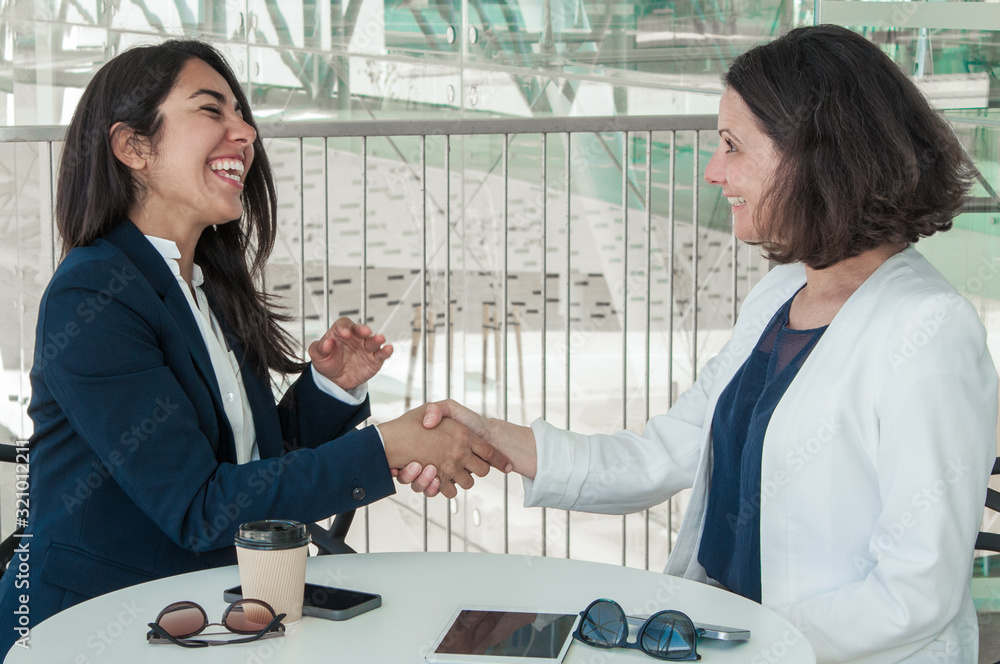 Fototapeta premium Two happy business women shaking hands in modern cafe. Partners sitting at table with tablet, smartphones and disposable cup. Meeting and agreement concept. Side view.