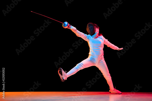 Forward. Teen girl in fencing costume with sword in hand isolated on black background, neon light. Young model practicing and training in motion, action. Copyspace. Sport, youth, healthy lifestyle.