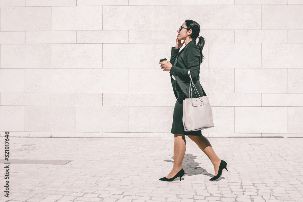 Serious professional late for work an drinking takeaway coffee on run. Young business woman in formal suit walking outdoors, carrying handbag and takeaway coffee. Morning way to office concept Serious professional late for work an drinking takeaway coffee on run. Young business woman in formal suit walking outdoors, carrying handbag and takeaway coffee. Morning way to office concept