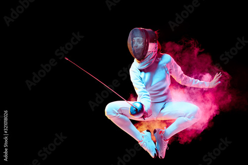 Pride. Teen girl in fencing costume with sword in hand isolated on black background, neon lighted smoke. Practicing and training in motion, action. Copyspace. Sport, youth, healthy lifestyle.