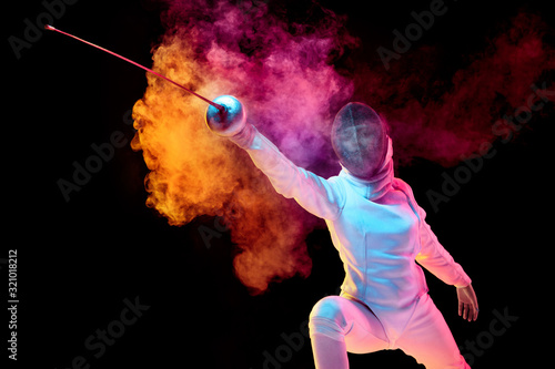 Contrasts. Teen girl in fencing costume with sword in hand isolated on black background, neon lighted smoke. Practicing and training in motion, action. Copyspace. Sport, youth, healthy lifestyle. photo