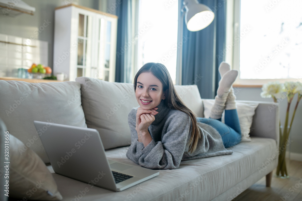 Young girl in a grey sweater laying on the sofa and chatting online Young girl in a grey sweater laying on the sofa and chatting online