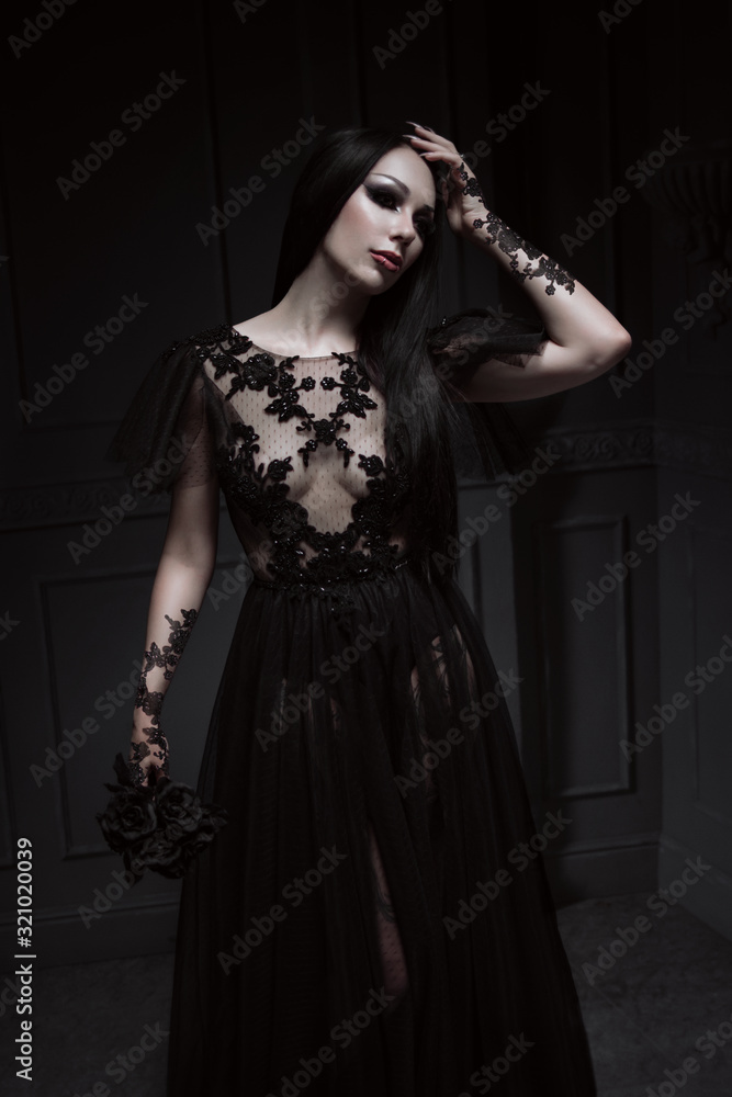 Beautiful brunette woman with gothic makeup wearing stylish black dress and posing on grey background