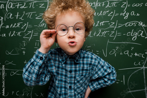 smart kid touching glasses and standing with hand on hip near chalkboard photo