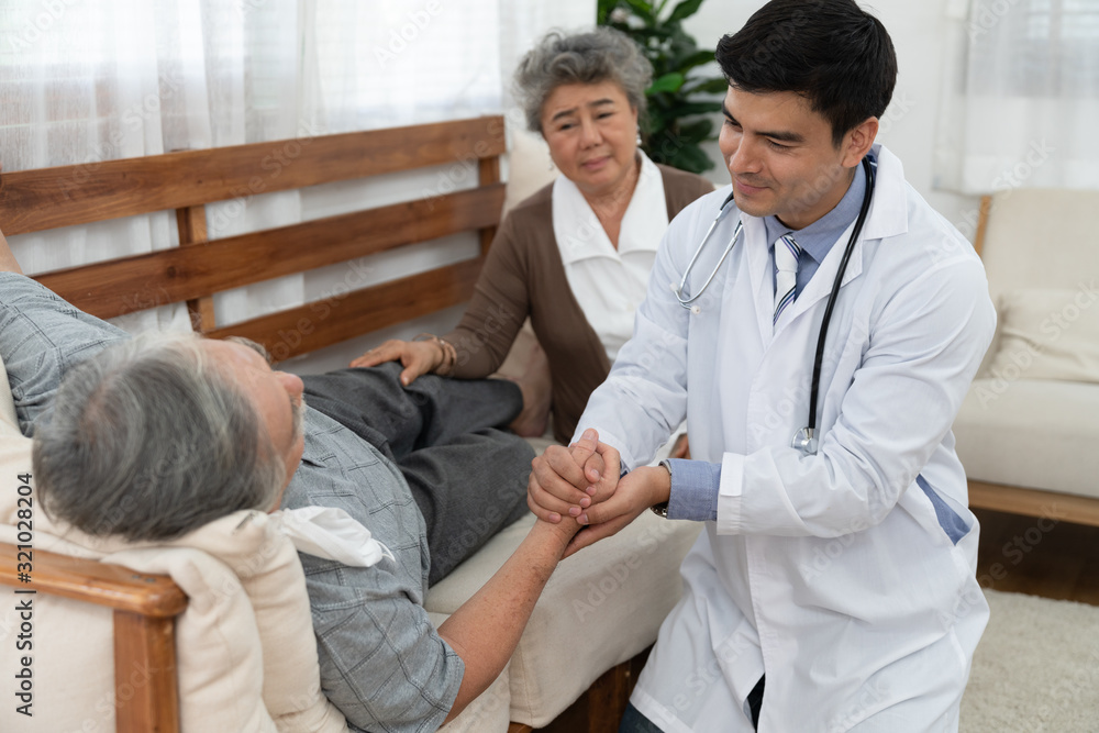 Doctor holding hand elderly man patient lying on sofa with his wife sitting near on his side after doctor take medical examination and friendly encouragement Doctor holding hand elderly man patient lying on sofa with his wife sitting near on his side after doctor take medical examination and friendly encouragement