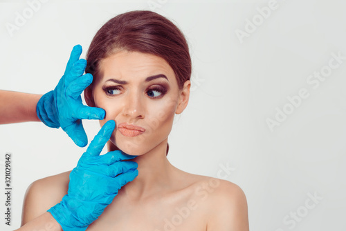 Preparing for cosmetic plastic surgery . Skeptical worried woman looks to the surgeon hands while the doctor examines her nasolabial wrinkle crease doubtful scared afraid, isolated white background photo