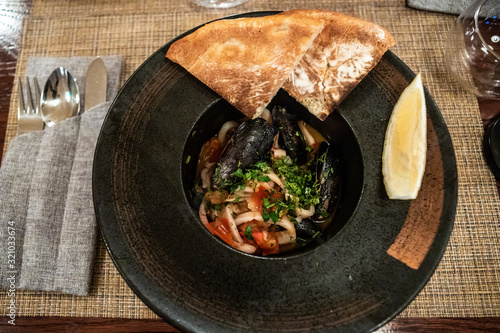 Roast mussels and squid with tomatoes and herbs in a dark plate with lemon and slices of bread. Delicacy