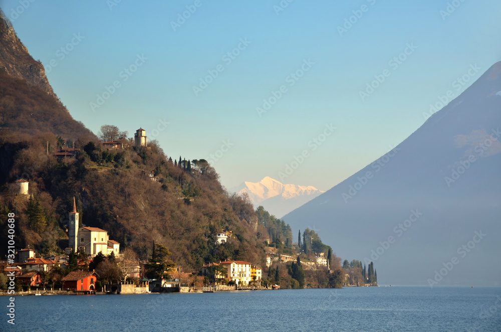 Sunlight over an Alpine Lake and Village with Snow-capped Mountain in Lombardy, Italy.
