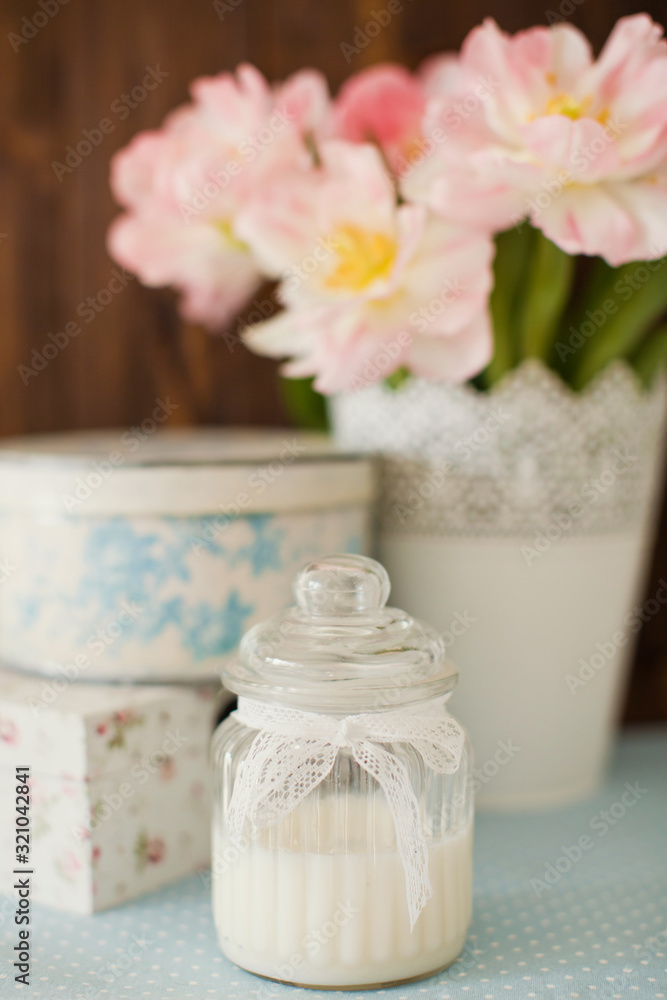White natural candle in vintage glass jar with lace ribbon. Gift boxes with flower ornament. Bouquet of tulips with pink and white petals in white metal vase.