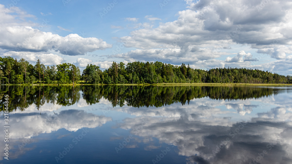 Wonderful landscape with lake on a sunny summer day. Blue sky with cumulus clouds, forest on the other side reflected in calm water. Latvia.
