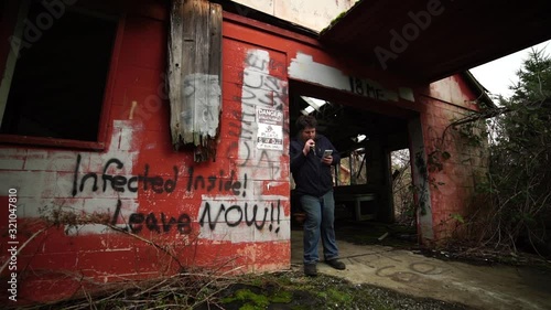 A young Millennial man leaning in the dorrway of an abondoned building scolling on his cell phone and vaping, slow motion photo