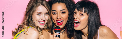 panoramic shot of attractive happy multiethnic girls singing karaoke with microphone, isolated on pink