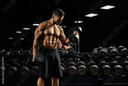 Shirtless man building biceps with dumbbell in gym.