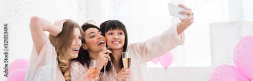 panoramic shot of emotional multicultural girls with glasses of champagne taking selfie on smartphone during pajama party