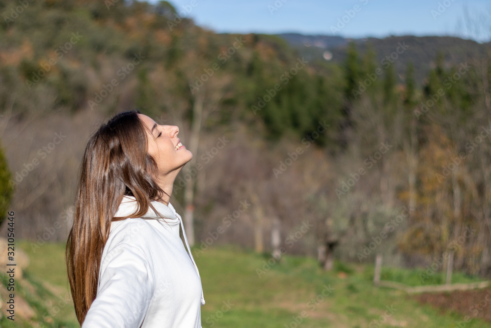 Smiling pretty young woman with sweatshirt breathing clean air in nature and sunbathing and mountains behind Smiling pretty young woman with sweatshirt breathing clean air in nature and sunbathing and mountains behind