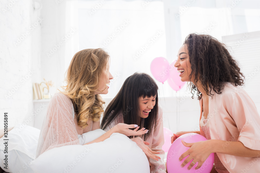 happy multicultural girlfriends laughing on bed with pink balloons happy multicultural girlfriends laughing on bed with pink balloons
