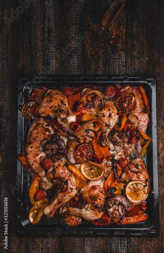 Roasted chicken legs with various vegetables and oranges on metal tray, wooden background, top view