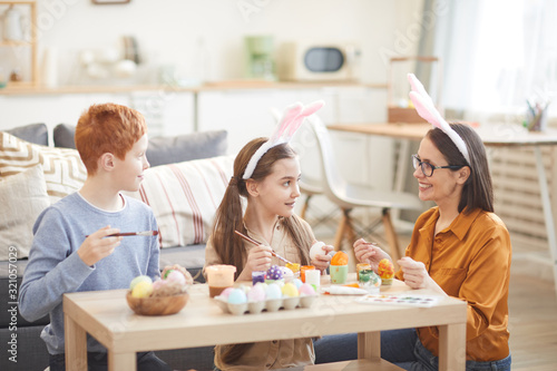 Young smiling mother painting Easter eggs together with her children at the table in the living room at home