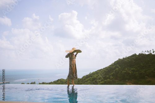 A beautiful girl in a swimsuit, a hat and a robe developing in the wind stands in a villa with a pool and beautiful views of the jungle and the sea. Thailand, Samui