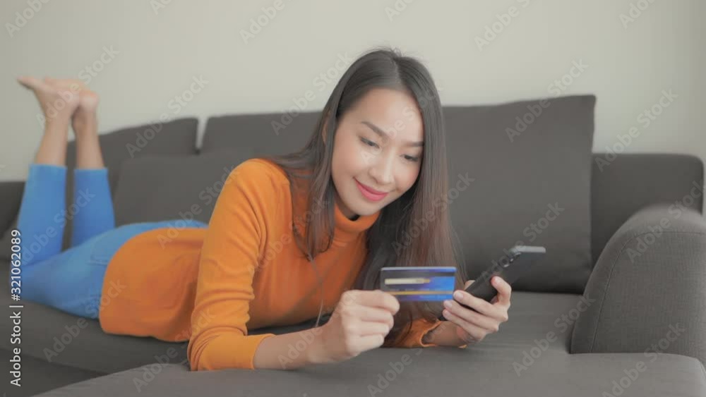 Close-up of a young woman lying on her stomach on a couch inputting her credit card number into her smartphone. Close-up of a young woman lying on her stomach on a couch inputting her credit card number into her smartphone.