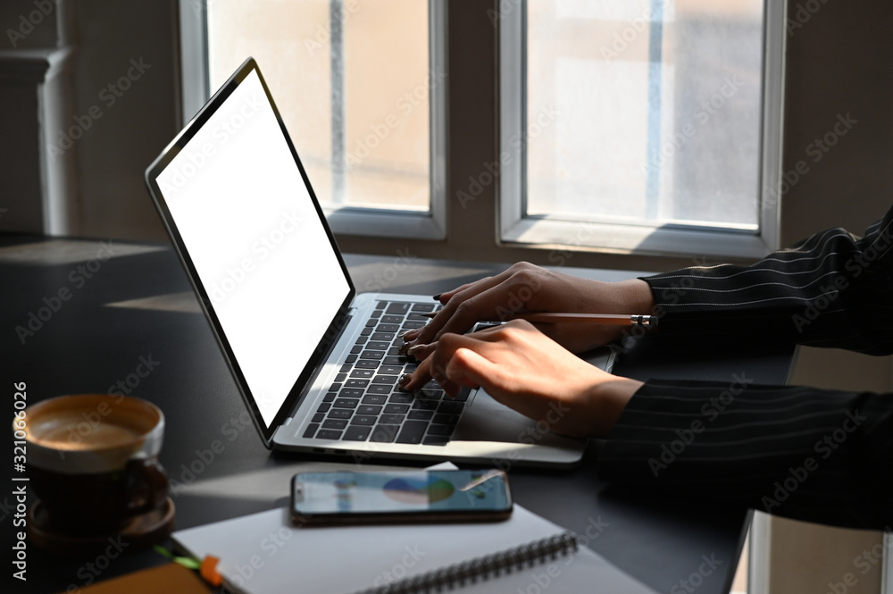 Side shot of young businesswoman typing on white blank screen laptop while sitting at the modern working desk. Coffee cup, smartphone with graphic chart on display putting together on table. Side shot of young businesswoman typing on white blank screen laptop while sitting at the modern working desk. Coffee cup, smartphone with graphic chart on display putting together on table.