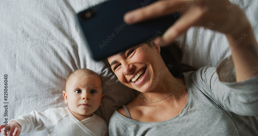 Authentic close up of neo mother and her newborn baby making a selfie or video call to father or relatives in a bed. Concept of technology, new generation,family, connection, parenthood, authenticity - obrazy, fototapety, plakaty 