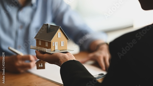 Cropped shot of House broker holding the sample house model in hand with his customer while writing on paper/ signing an agreement at the wooden table. Loan,Debt, selling/buying agreement concept.