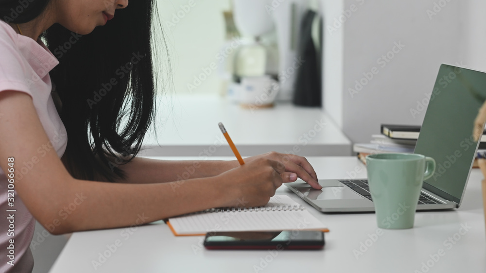 Cropped shot of young beautiful woman writing/taking notes while sitting at the modern working desk with comfortable living room as background. Cropped shot of young beautiful woman writing/taking notes while sitting at the modern working desk with comfortable living room as background.