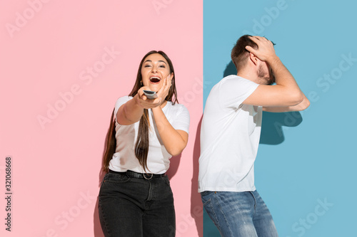 Who's in control. She chooses TV channel, he's angry. Young man, woman in casual on pink, blue bicolored background. Concept of human emotions, facial expession, relations, ad. Beautiful couple.
