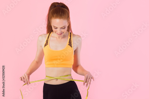beautiful young slim woman with red hair in sportswear on a pink background with a measuring tape © Alexandr
