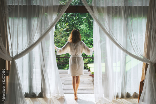 A woman in a bathrobe opens the curtains in deluxe Bali hotel room overlooking the terrace and tropical trees.Woman is awoke and standing before window. Girl is opening curtains and meeting sunrise