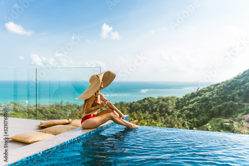 A beautiful girl in a red bathing suit is relaxing in a luxury villa by the pool with wine and in a hat taking cover from the sun. View of palm trees and the sea. Thailand, Koh Samui © Underwater girls