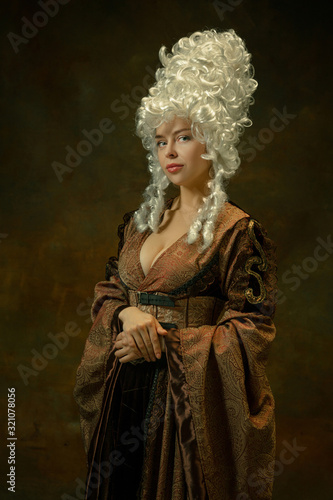 Portrait of medieval young woman in brown vintage clothing standing on dark background. Female model as a duchess, royal person. Concept of comparison of eras, modern, fashion, beauty.