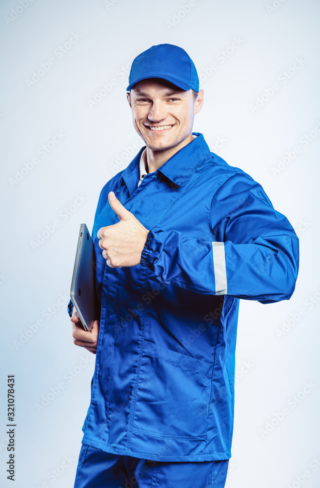 Portrait of young worker man wearing blue uniform. Holding a laptop in his hand and showing thumb up. Movement cool. Isolated on grey background with copy space. Business concept. Portrait of young worker man wearing blue uniform. Holding a laptop in his hand and showing thumb up. Movement cool. Isolated on grey background with copy space. Business concept.